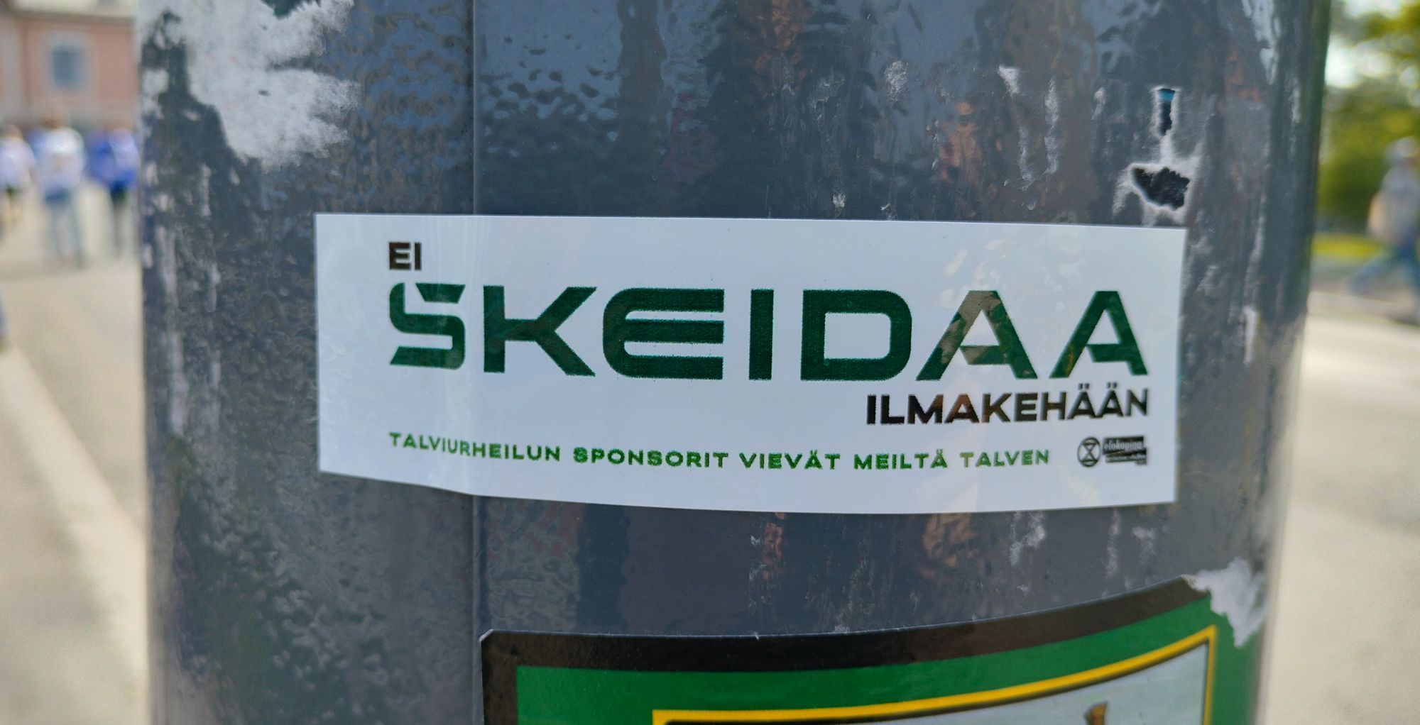 Metallipintaan liimattu tarra jossa teksti: Ei skeidaa ilmakehään – talviurheilun sponsorit vievät meiltä talven + EK-logo.